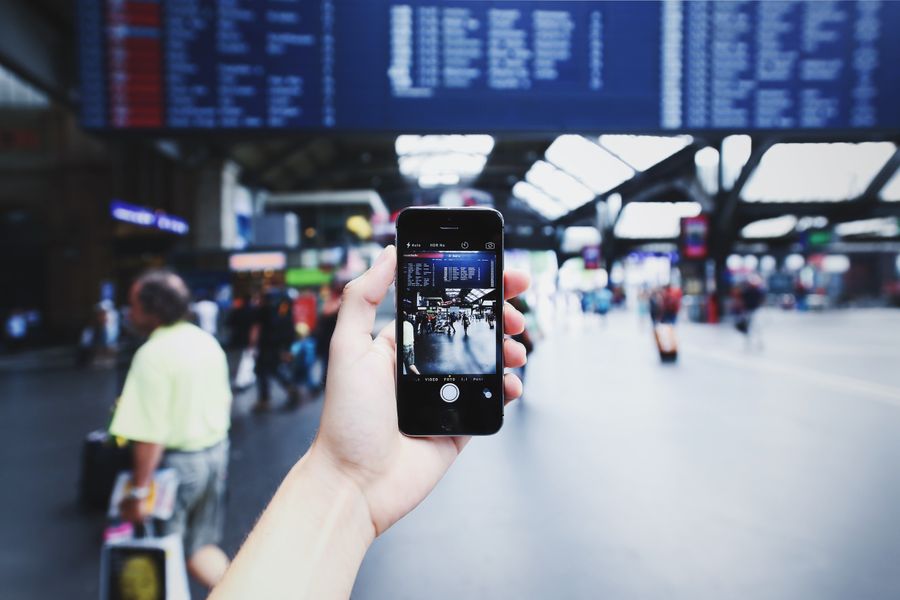 A hand holding a smartphone capturing a bustling train station with blurred travelers in the background. — this tool: tell it about the trip, get the answer in under a minute.
