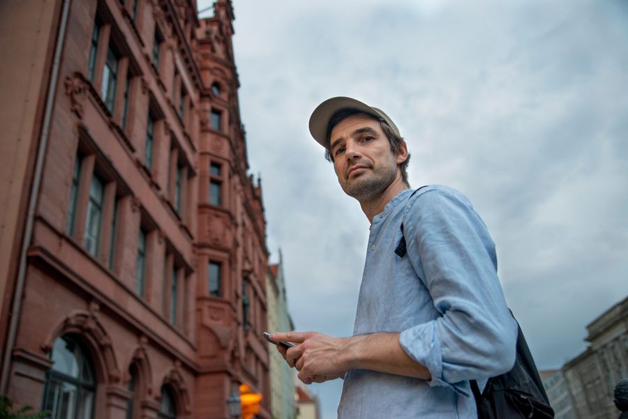 Casually dressed man using smartphone in Berlin city street with historic architecture. — Sprache oder Text: Sprechen Sie über Abdeckung, Tarife und Troubleshooting.