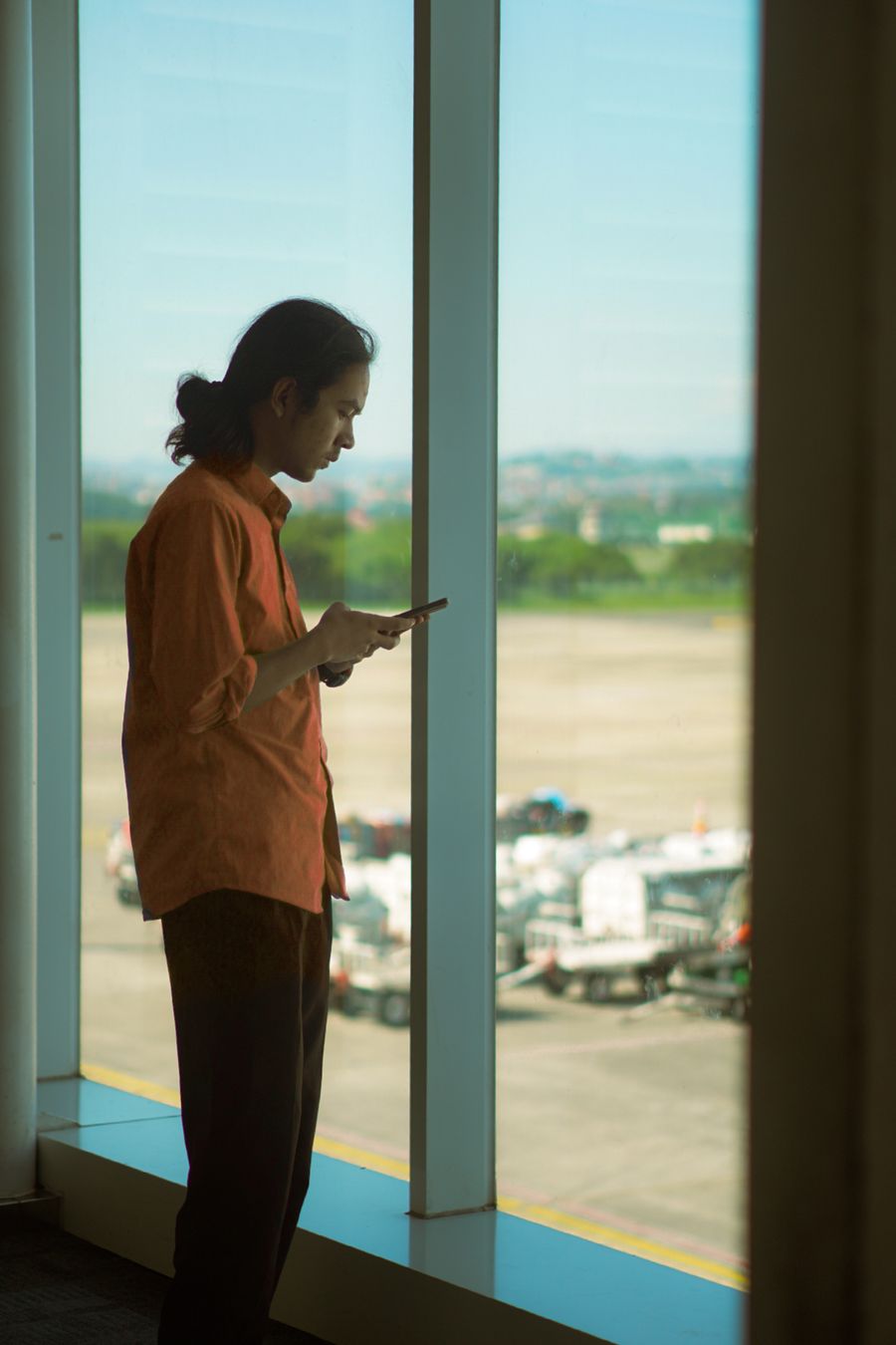 Man in orange shirt using a smartphone while standing at an airport window, overlooking the runway. — The rental desk handshake: confirmation email open, license photo ready.