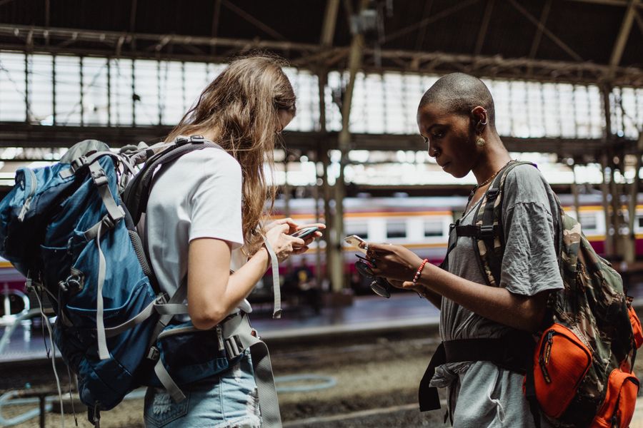 Two adventurous backpackers using smartphones while waiting at a bustling train station. — Kein Ladenbesuch, keine Kioskjagd, keine Passkopien.