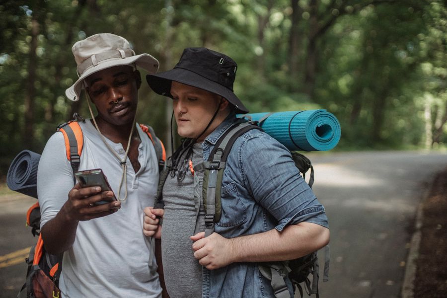 Young multiethnic tourists with hiking equipment surfing internet on cellphone on roadway against trees. — The night bus: entertainment downloaded, messages queued, rest easy.